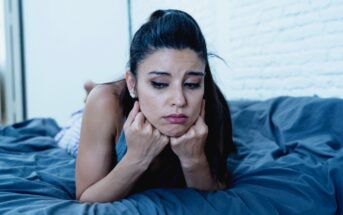 A woman with long dark hair lies on a bed, resting her chin on her fists. She has a pensive, slightly sad expression. The bedding is dark blue, and there is a white brick wall in the background.