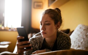 A woman with a bun hairstyle is sitting on a sofa, holding and looking at a smartphone. The background is softly lit, with a window partially visible, creating a cozy atmosphere in the room.