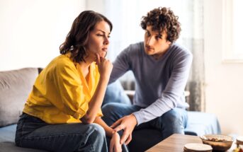 A woman in a yellow shirt sits on a couch with a thoughtful expression, resting her chin on her hand. A man in a blue sweater sits beside her, looking at her with concern. They are in a cozy living room setting.