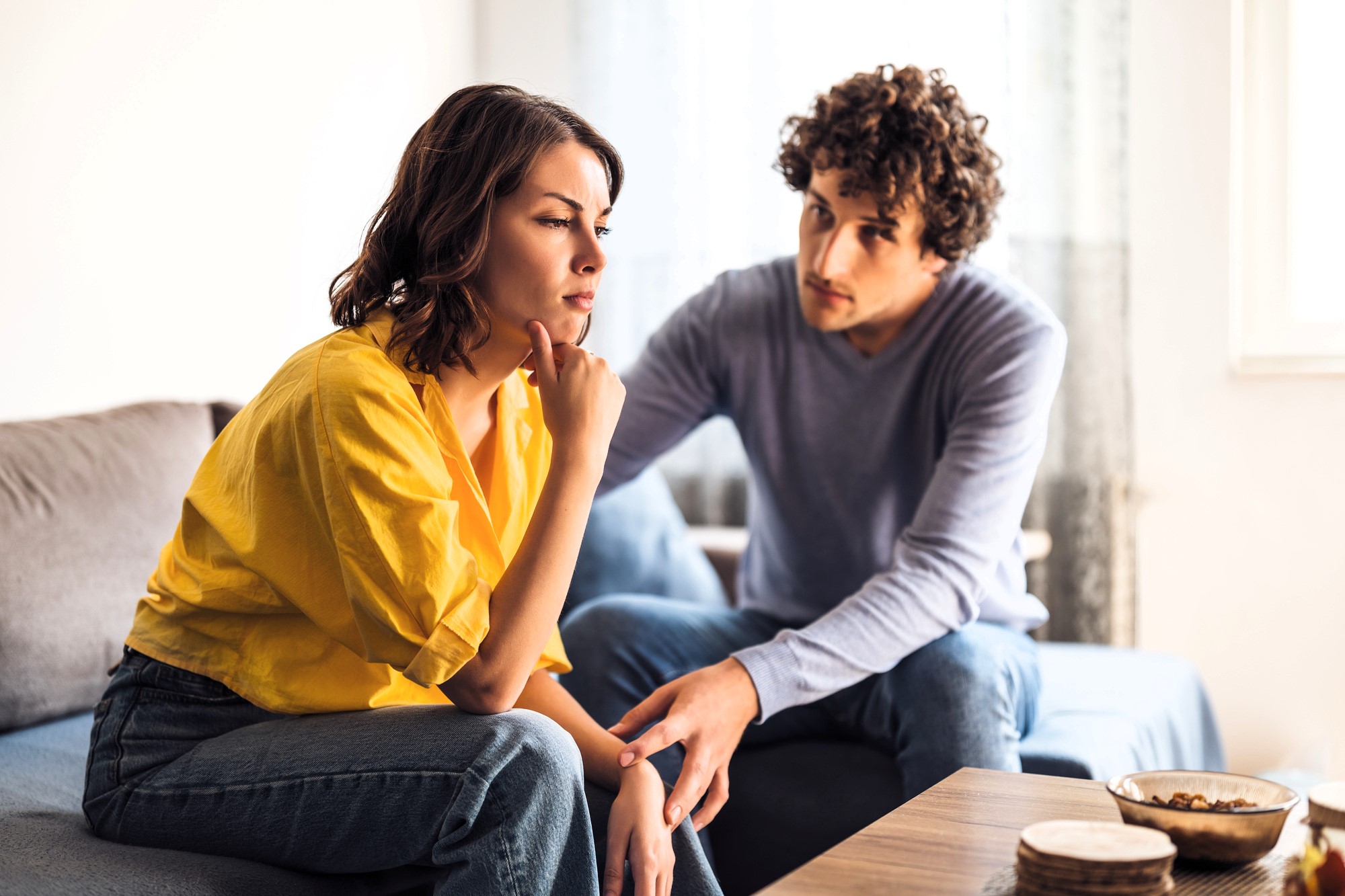 A woman in a yellow shirt sits on a couch with a thoughtful expression, resting her chin on her hand. A man in a blue sweater sits beside her, looking at her with concern. They are in a cozy living room setting.