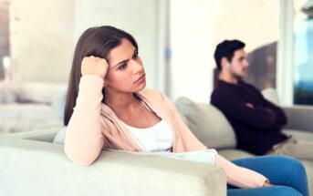 A woman with long brown hair, wearing a pink cardigan, sits on a couch with her head propped on her hand, looking pensive. In the background, a man sits on another couch with crossed arms, facing away. They appear to be in a living room.