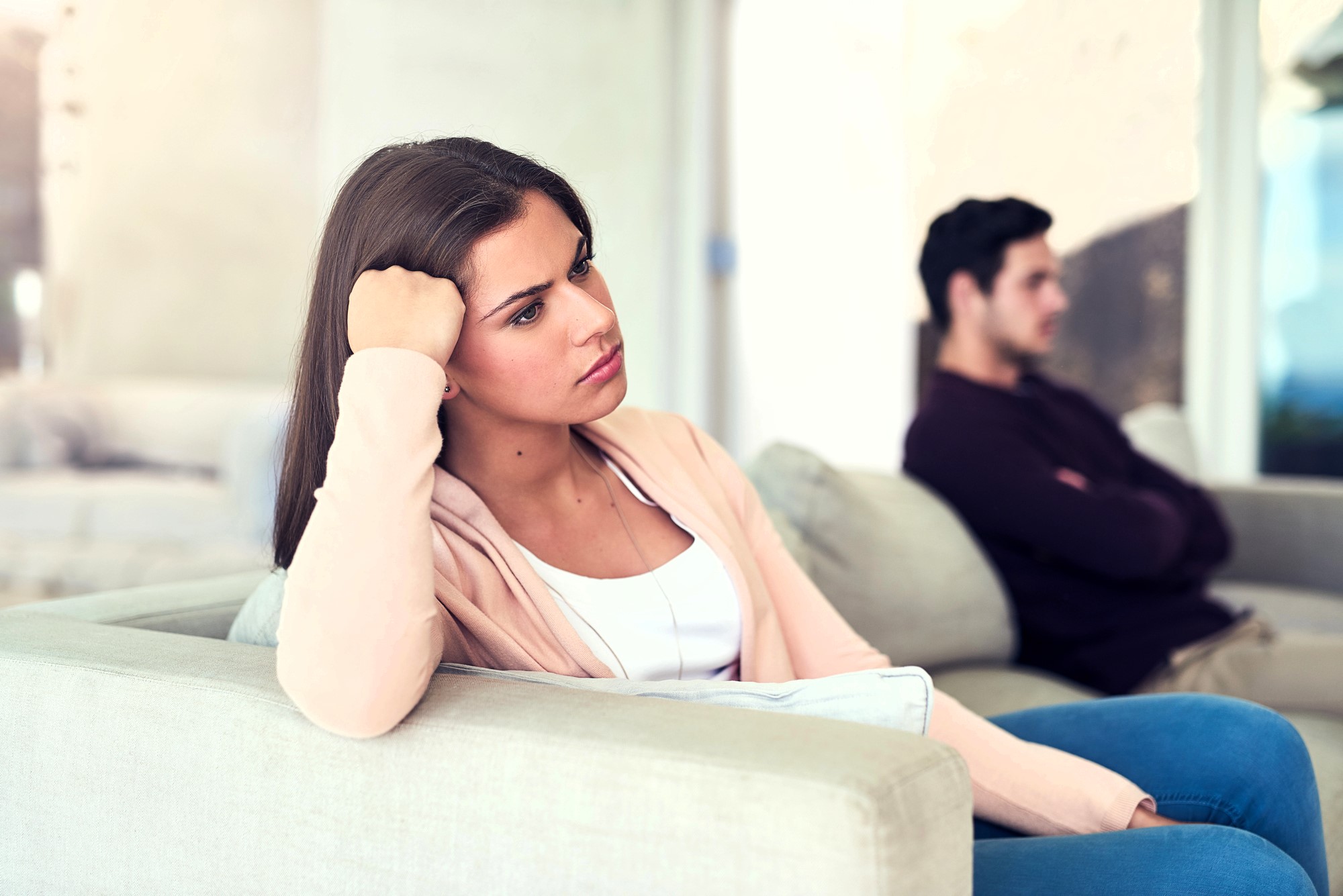 A woman with long brown hair, wearing a pink cardigan, sits on a couch with her head propped on her hand, looking pensive. In the background, a man sits on another couch with crossed arms, facing away. They appear to be in a living room.