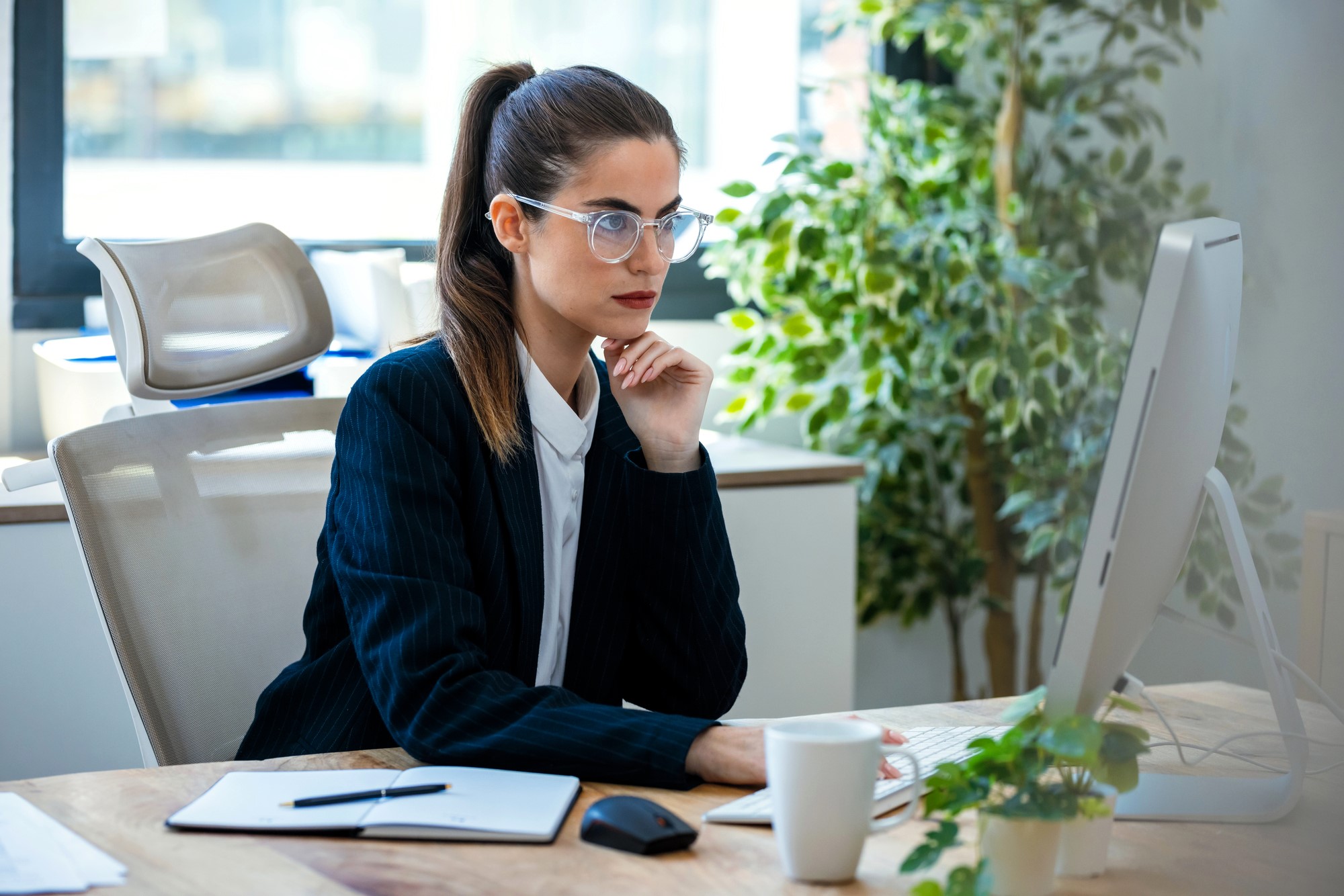 A woman in a suit and glasses works at a computer in a modern office. She appears focused, with her chin resting on her hand. The desk has a notebook, a coffee cup, and a potted plant. Large windows and greenery are in the background.