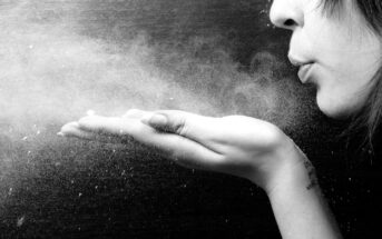 Black and white image of a person blowing powder from their hand. The fine particles create a misty effect against a dark background. The person's face is partially visible, showing pursed lips.