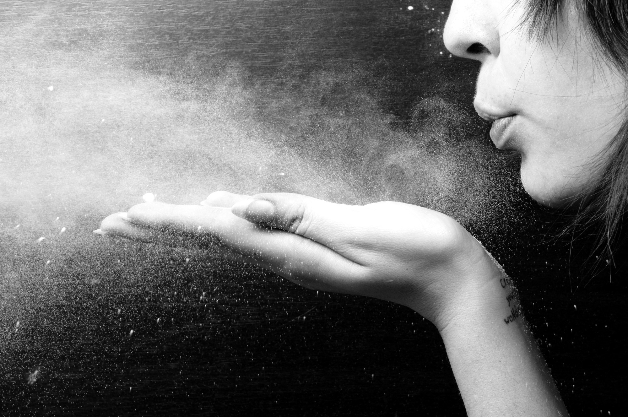 Black and white image of a person blowing powder from their hand. The fine particles create a misty effect against a dark background. The person's face is partially visible, showing pursed lips.