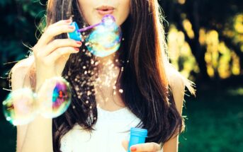 A woman with long brown hair is blowing bubbles outdoors. She holds a blue bubble wand and container, with sunlight creating colorful reflections on the bubbles. The background is blurred greenery.