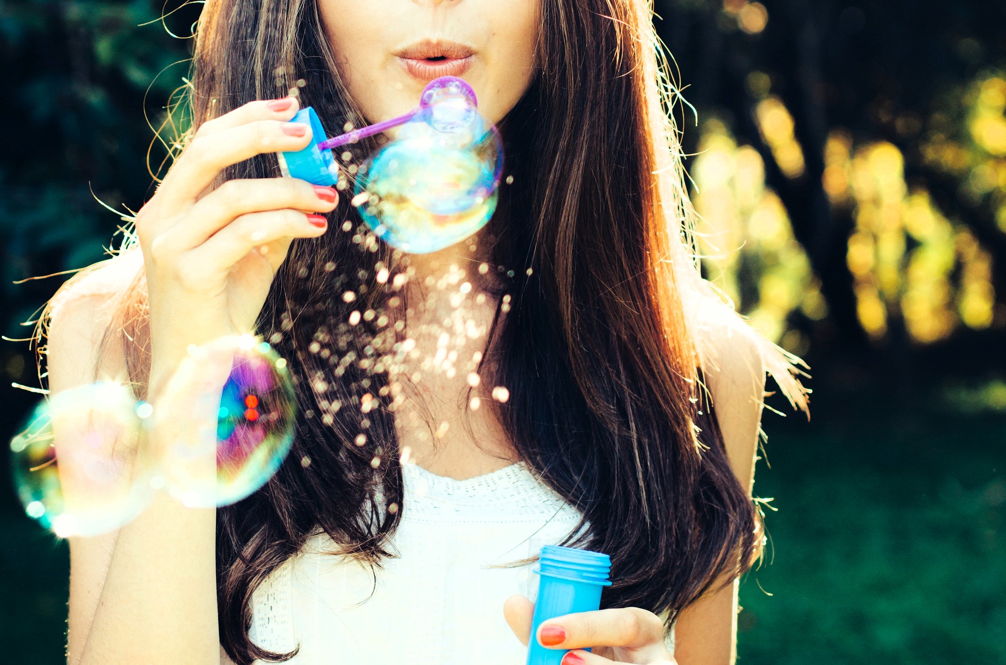 A woman with long brown hair is blowing bubbles outdoors. She holds a blue bubble wand and container, with sunlight creating colorful reflections on the bubbles. The background is blurred greenery.