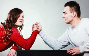 A woman in a red sweater and a man in a white sweater arm-wrestle across a table. Both are focused, with the woman showing a determined expression. The background is plain gray.