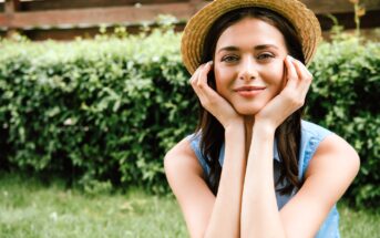 Woman wearing a straw hat, resting her chin on her hands and smiling, is sitting outside with a green hedge in the background. She wears a blue sleeveless top.