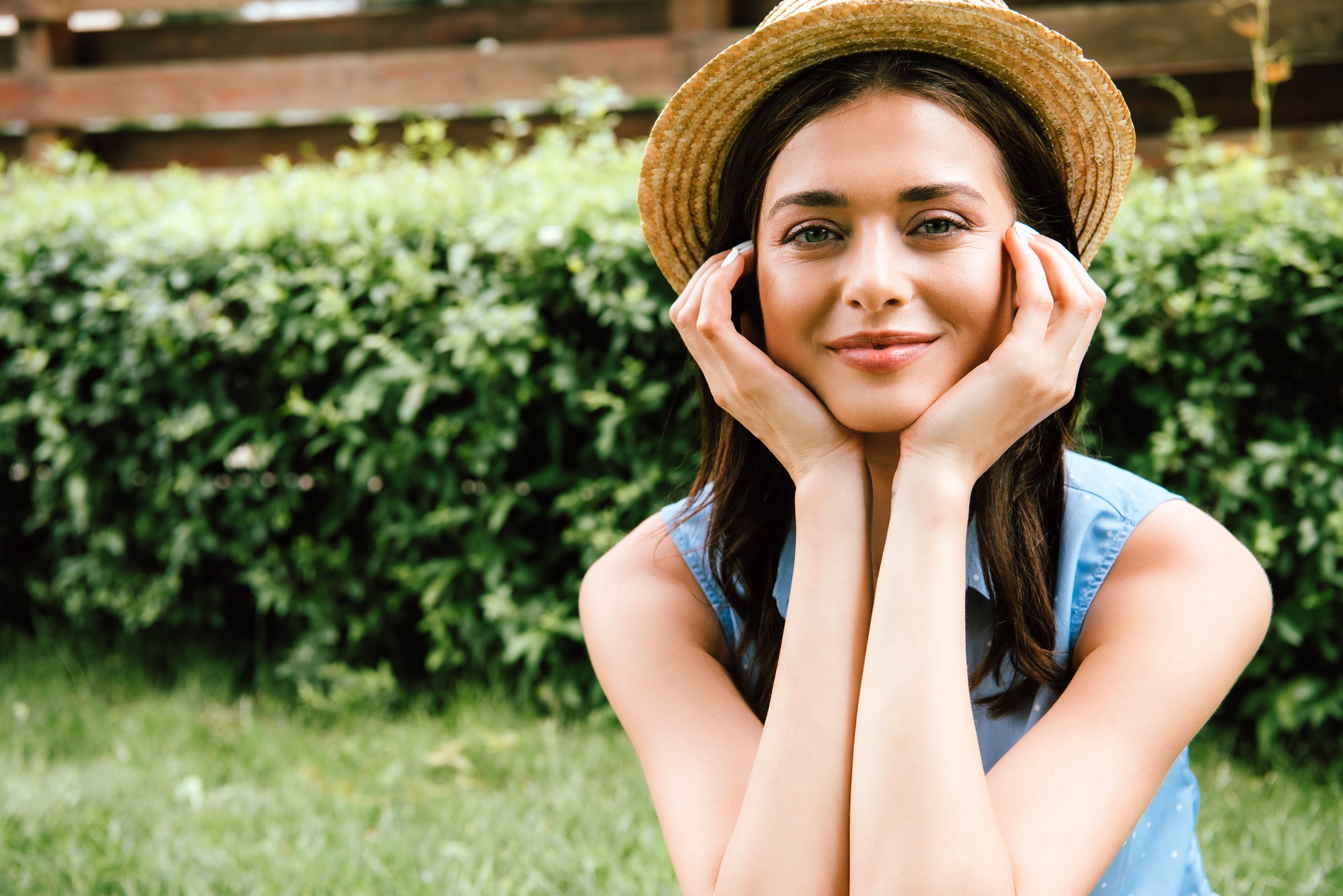 Woman wearing a straw hat, resting her chin on her hands and smiling, is sitting outside with a green hedge in the background. She wears a blue sleeveless top.