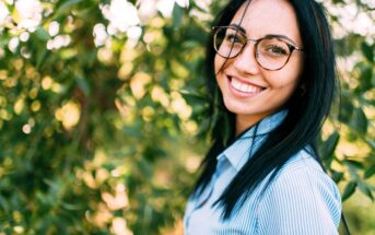 A smiling woman with long black hair and glasses stands outdoors in front of green foliage. She is wearing a light blue and white striped shirt. The sunlight filters through the leaves in the background.