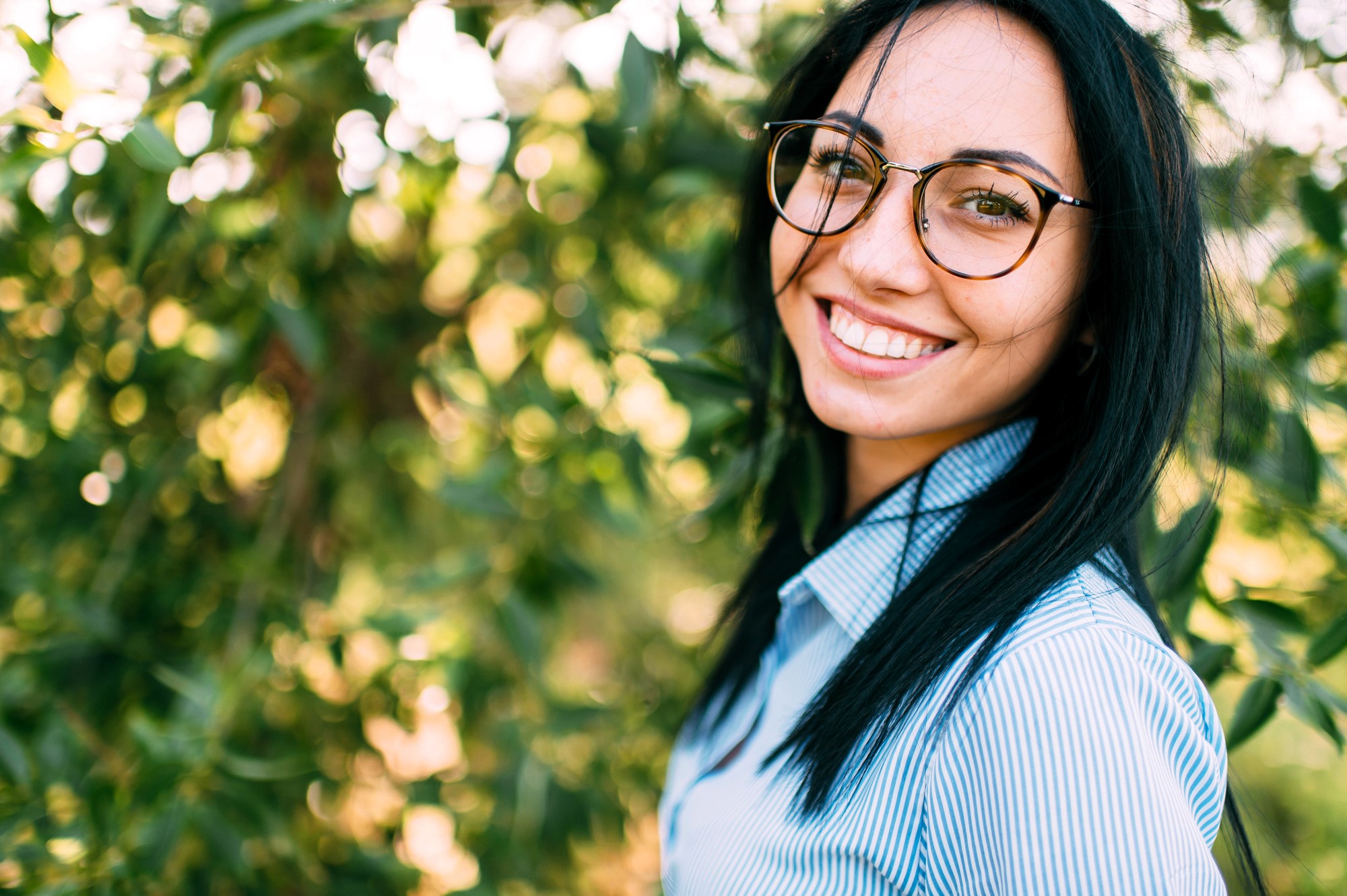 A smiling woman with long black hair and glasses stands outdoors in front of green foliage. She is wearing a light blue and white striped shirt. The sunlight filters through the leaves in the background.