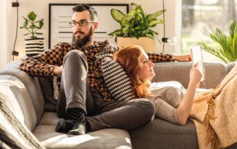 A bearded man wearing glasses and a plaid shirt relaxes on a couch with a woman reading a book, leaning against his leg. They are surrounded by decorative pillows and plants, with sunlight streaming in through a nearby window.