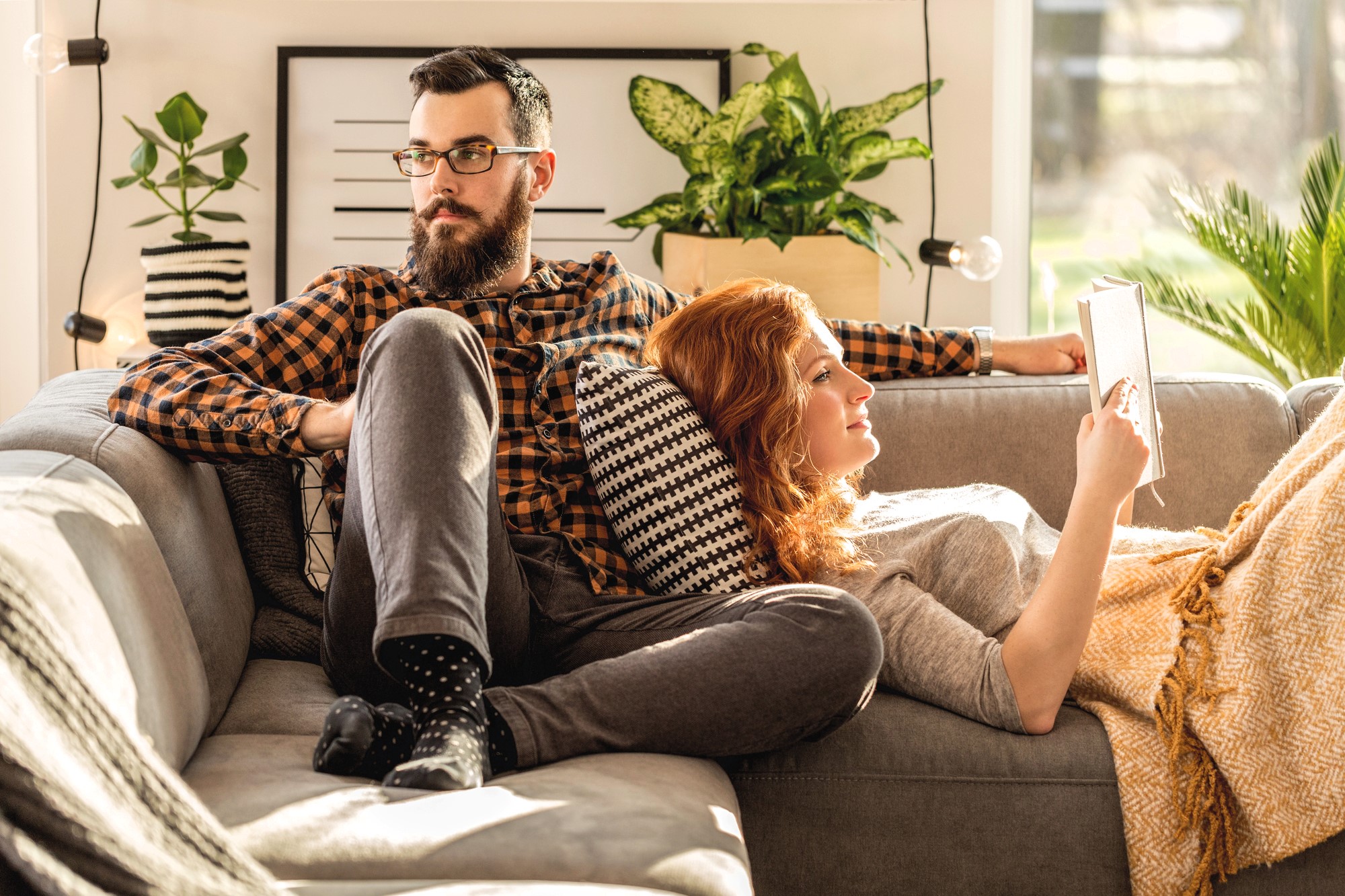 A bearded man wearing glasses and a plaid shirt relaxes on a couch with a woman reading a book, leaning against his leg. They are surrounded by decorative pillows and plants, with sunlight streaming in through a nearby window.