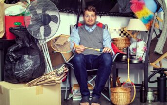 A man sitting in a cluttered space with various items around him, including a fan, badminton racket, wicker basket, colorful feather duster, and cardboard boxes. He wears a blue shirt, gray vest, and looks at the camera.