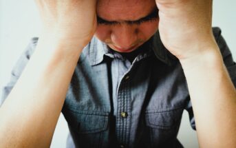 A person wearing a denim shirt holds their head in their hands, appearing stressed or deep in thought. The image is focused on the upper body and face, with a plain background.