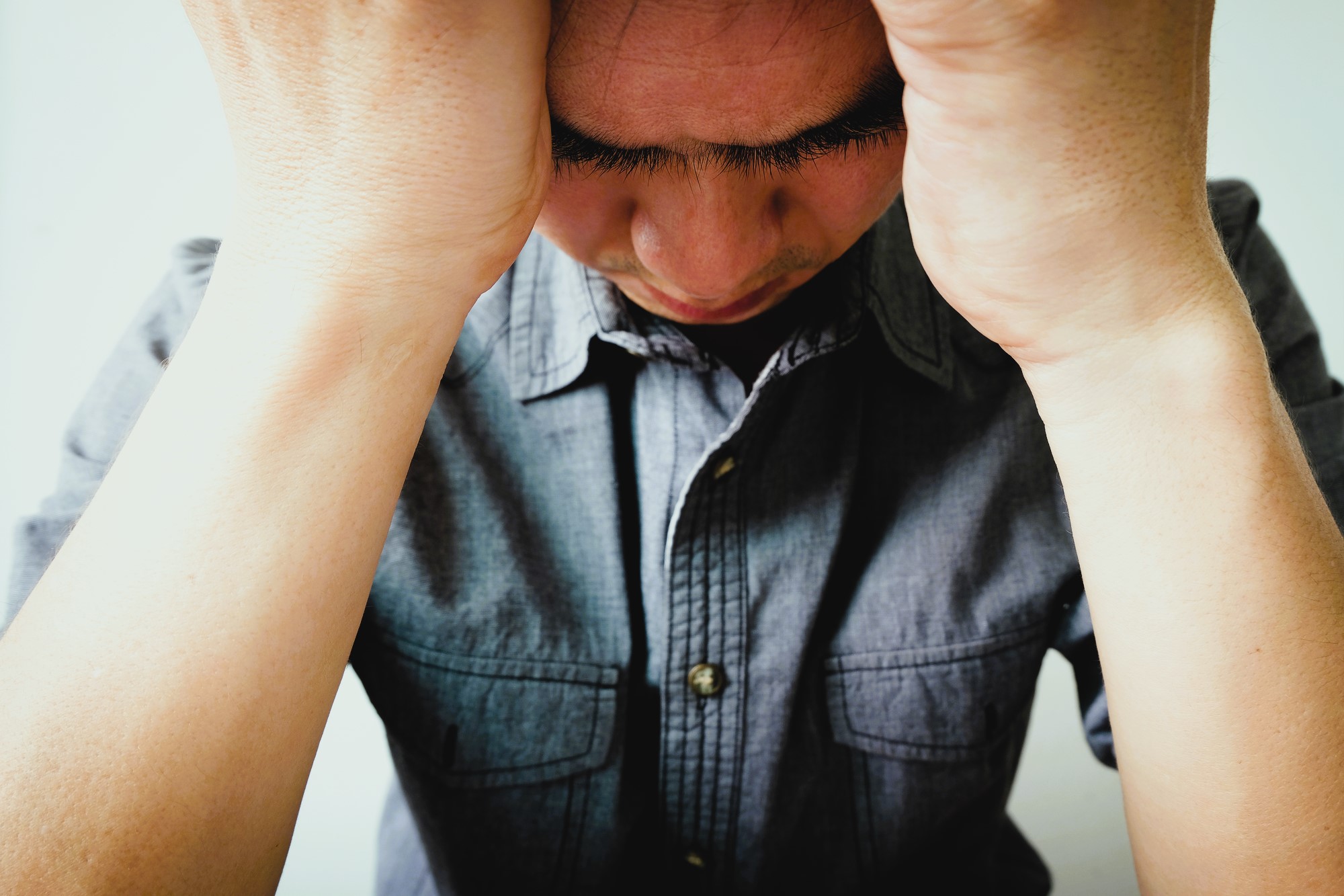 A person wearing a denim shirt holds their head in their hands, appearing stressed or deep in thought. The image is focused on the upper body and face, with a plain background.