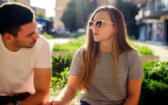 A young woman with long hair and sunglasses, wearing a striped shirt, talks animatedly to a man in a white t-shirt. They are sitting outside with a sunny background and greenery around them.