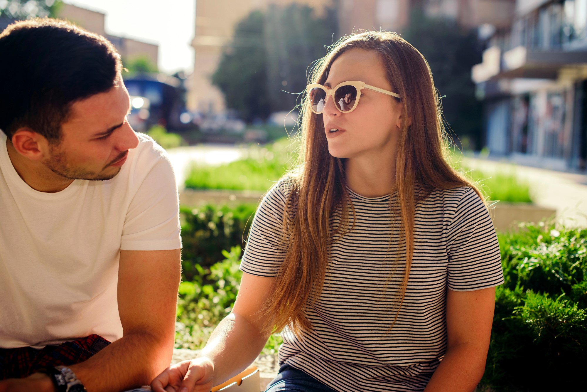 A young woman with long hair and sunglasses, wearing a striped shirt, talks animatedly to a man in a white t-shirt. They are sitting outside with a sunny background and greenery around them.