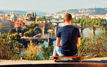 A person wearing a blue shirt and shorts sits on a skateboard, admiring a scenic view of a city with bridges over a river. The background shows lush greenery and buildings bathed in warm sunlight.