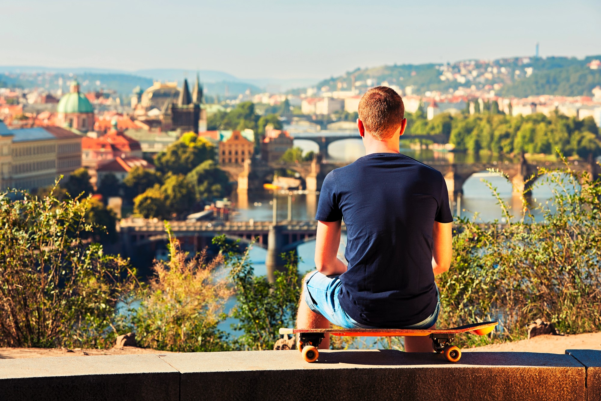 A person wearing a blue shirt and shorts sits on a skateboard, admiring a scenic view of a city with bridges over a river. The background shows lush greenery and buildings bathed in warm sunlight.