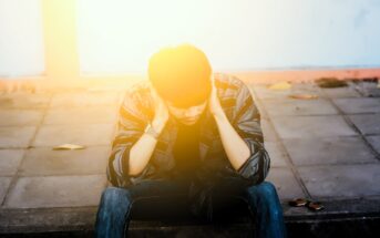 A person sits on a sidewalk with head in hands, under bright sunlight. They wear a striped shirt and jeans. The ground is paved with a few scattered leaves. The lighting creates a warm, dramatic effect.