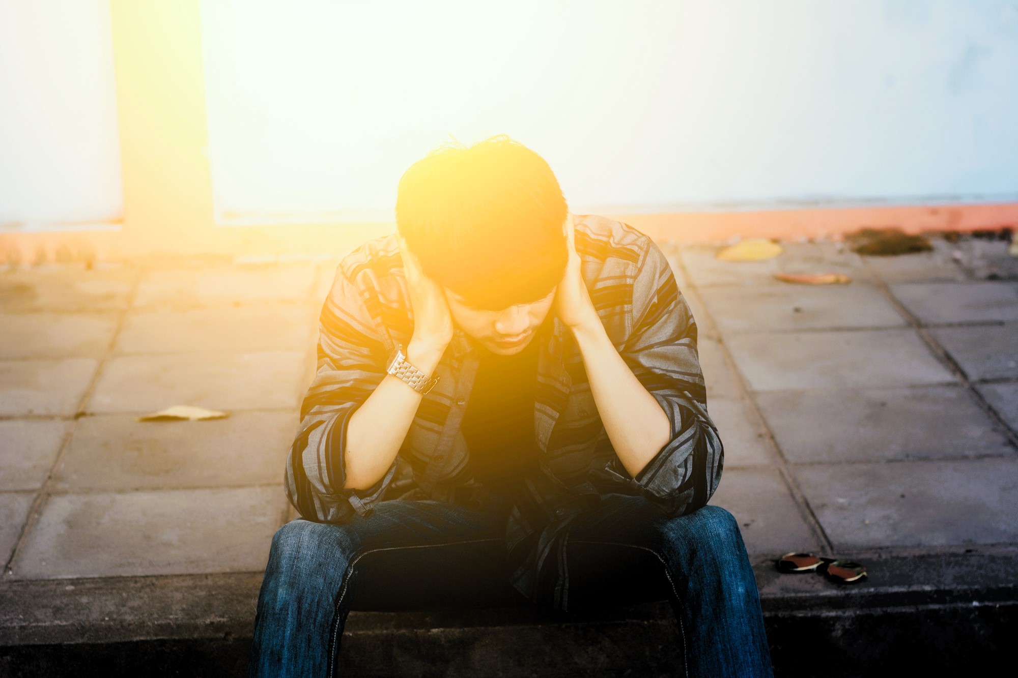 A person sits on a sidewalk with head in hands, under bright sunlight. They wear a striped shirt and jeans. The ground is paved with a few scattered leaves. The lighting creates a warm, dramatic effect.