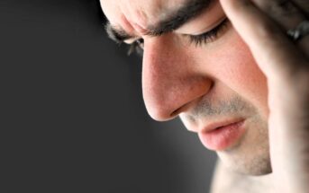 Close-up of a person with eyes closed and a pained expression, holding their head with one hand. The background is blurred, emphasizing the person's facial features and apparent discomfort.