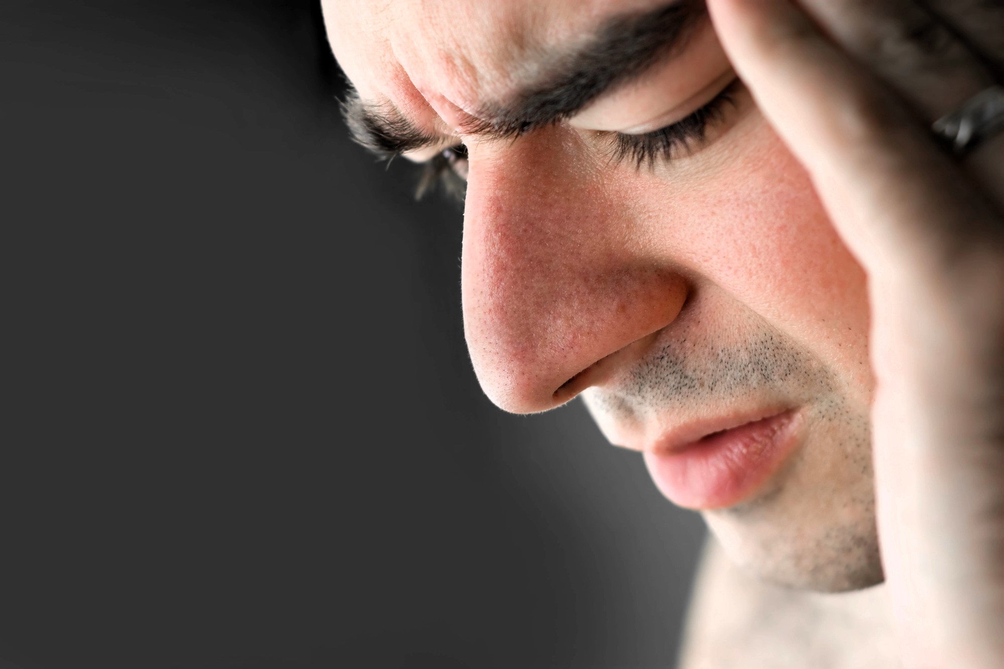 Close-up of a person with eyes closed and a pained expression, holding their head with one hand. The background is blurred, emphasizing the person's facial features and apparent discomfort.