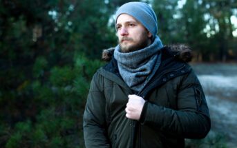 A bearded man wearing a gray beanie and scarf stands outside in a forested area. He is dressed in a green winter jacket with faux fur trim. He gazes thoughtfully into the distance, with trees blurred in the background.