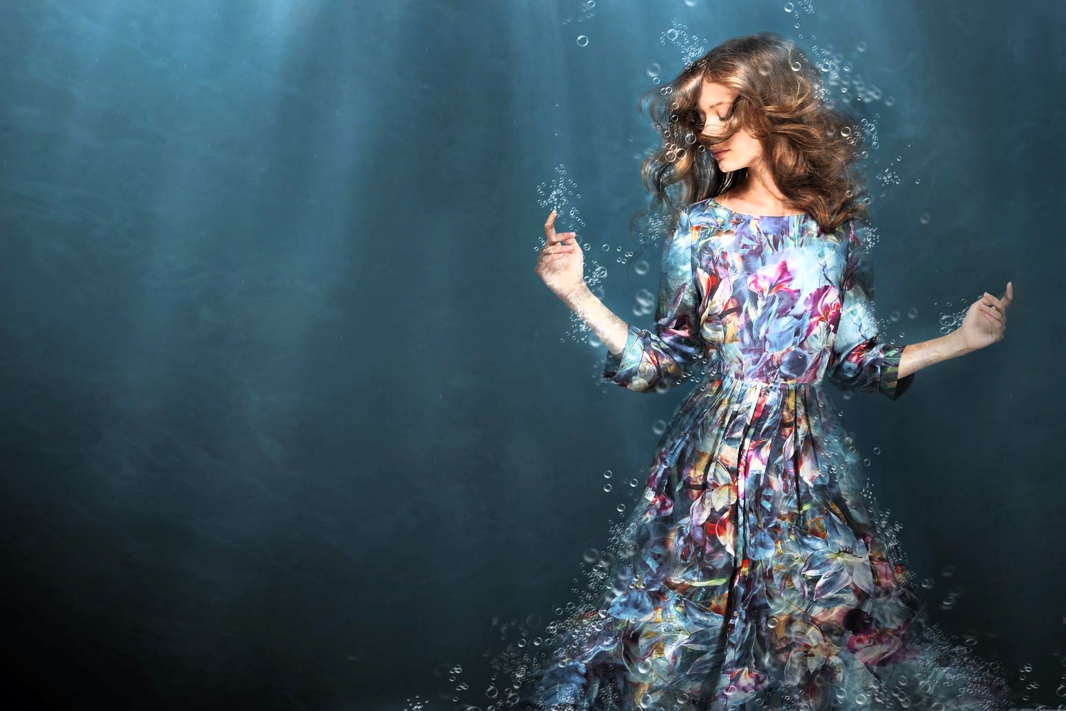 A woman with curly hair gracefully poses underwater, wearing a long, flowing dress with a colorful floral pattern. Bubbles surround her, and rays of light filter through the water, creating a serene atmosphere.