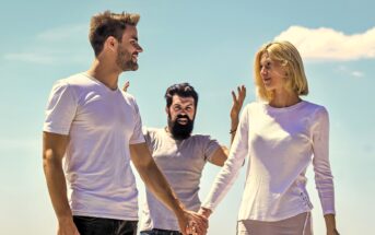 A couple smiling and holding hands, walking outdoors under a clear sky. A bearded man stands behind them with an excited expression, raising both arms in the air.