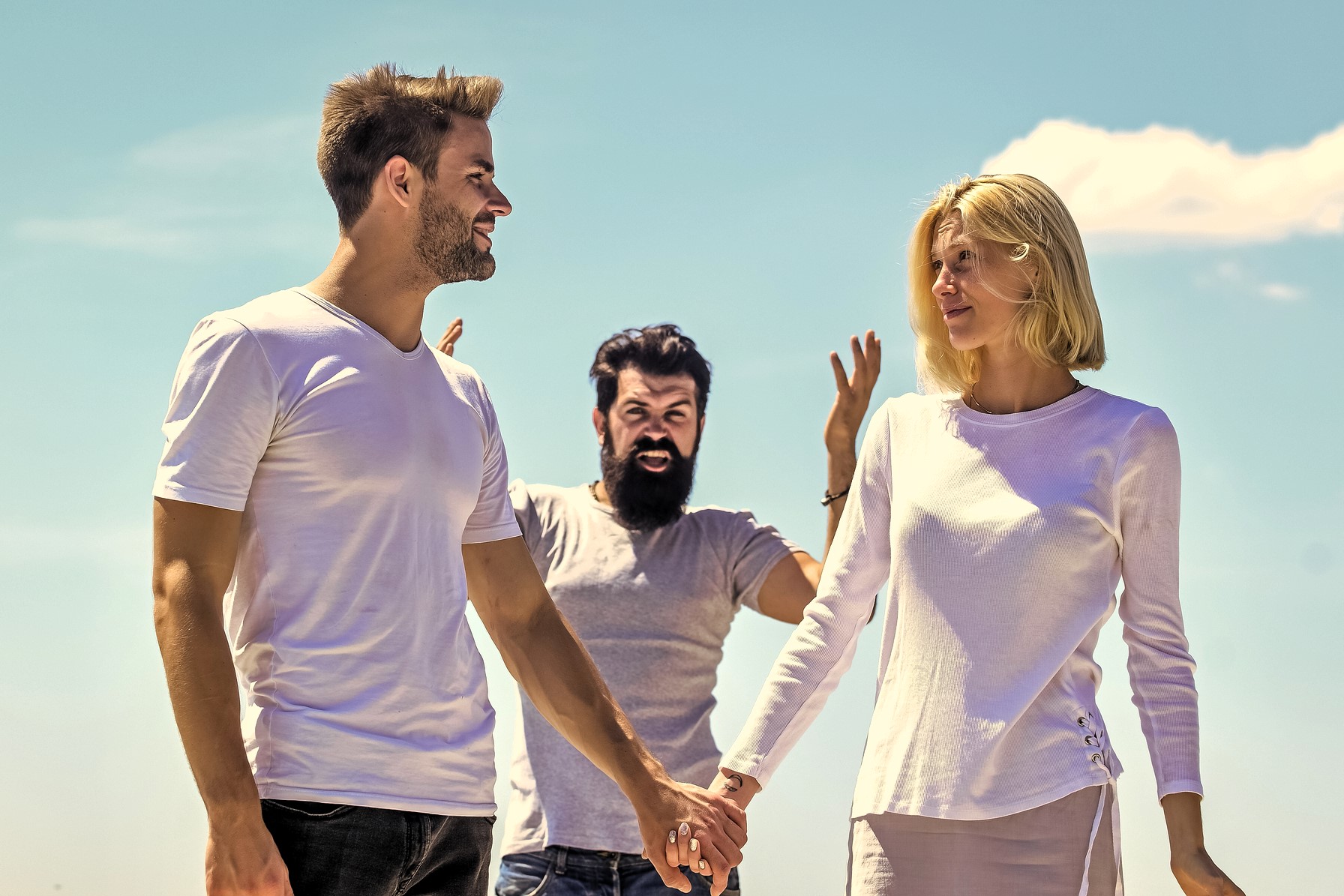 A couple smiling and holding hands, walking outdoors under a clear sky. A bearded man stands behind them with an excited expression, raising both arms in the air.