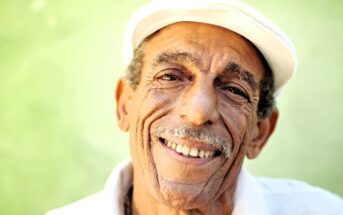 Elderly man with a white mustache smiles warmly, wearing a white cap and shirt. The green background adds a soft contrast to his tan complexion, enhancing the cheerful and welcoming expression on his face.