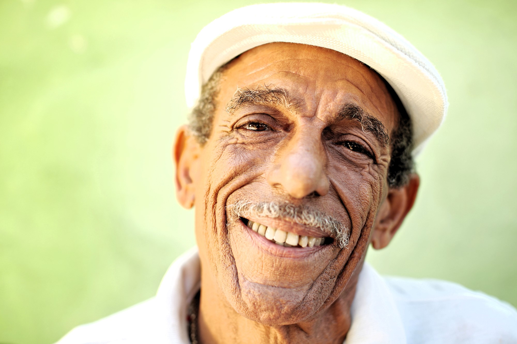 Elderly man with a white mustache smiles warmly, wearing a white cap and shirt. The green background adds a soft contrast to his tan complexion, enhancing the cheerful and welcoming expression on his face.