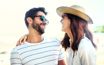 A smiling couple enjoying a sunny day outdoors. The man wears sunglasses and a striped shirt, while the woman has a straw hat and a white shirt. They are looking at each other with affection.