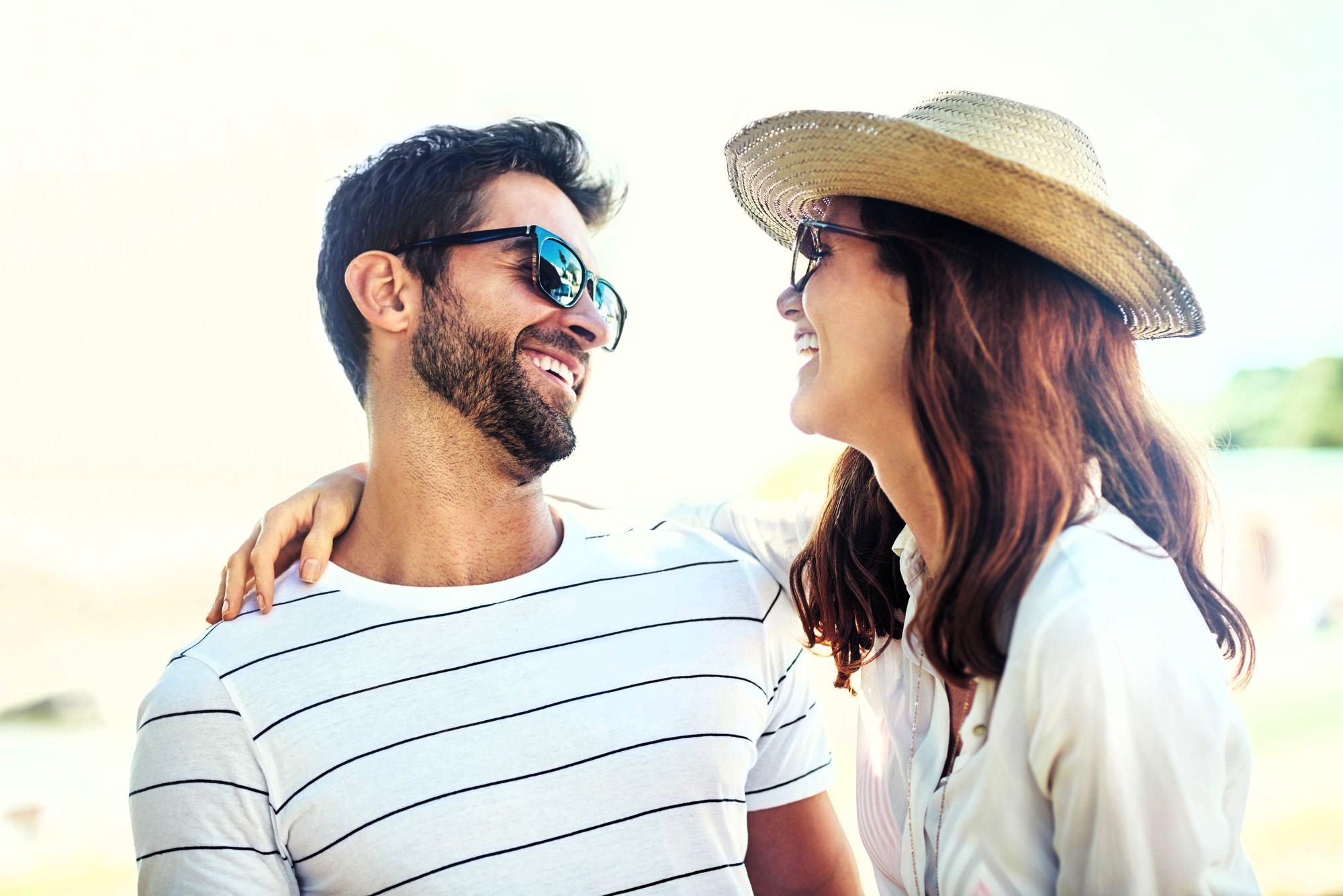 A smiling couple enjoying a sunny day outdoors. The man wears sunglasses and a striped shirt, while the woman has a straw hat and a white shirt. They are looking at each other with affection.