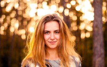 Woman with long hair smiling outdoors, standing in front of blurred trees and a sunlit background, which creates a bokeh effect.