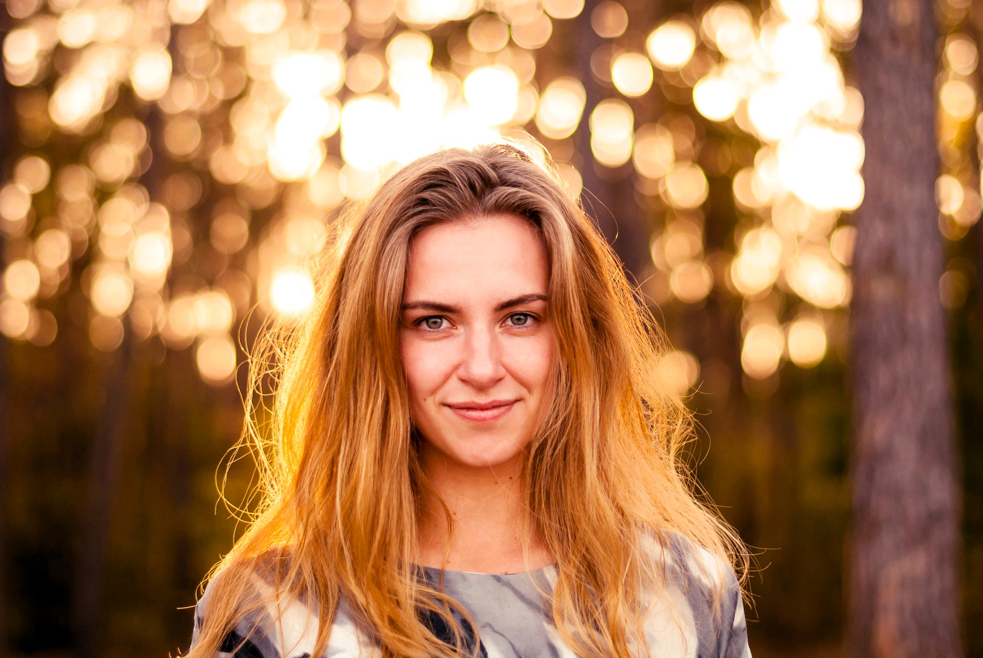 Woman with long hair smiling outdoors, standing in front of blurred trees and a sunlit background, which creates a bokeh effect.