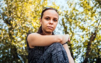 A woman with sunglasses on her head sits outdoors, arms resting on her knees. She wears a patterned top and gazes at the camera. The background is filled with blurred, sunlit trees, creating a warm, natural ambiance.