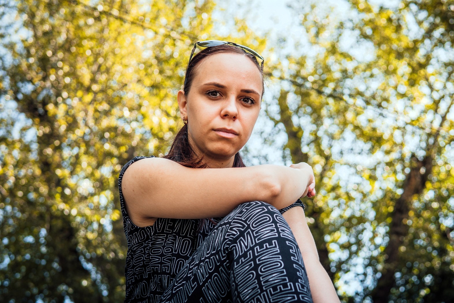 A woman with sunglasses on her head sits outdoors, arms resting on her knees. She wears a patterned top and gazes at the camera. The background is filled with blurred, sunlit trees, creating a warm, natural ambiance.