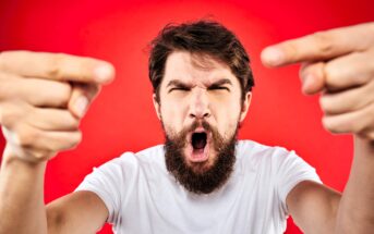 A bearded man in a white t-shirt, with an intense expression, points forcefully towards the camera. He is against a bright red background.