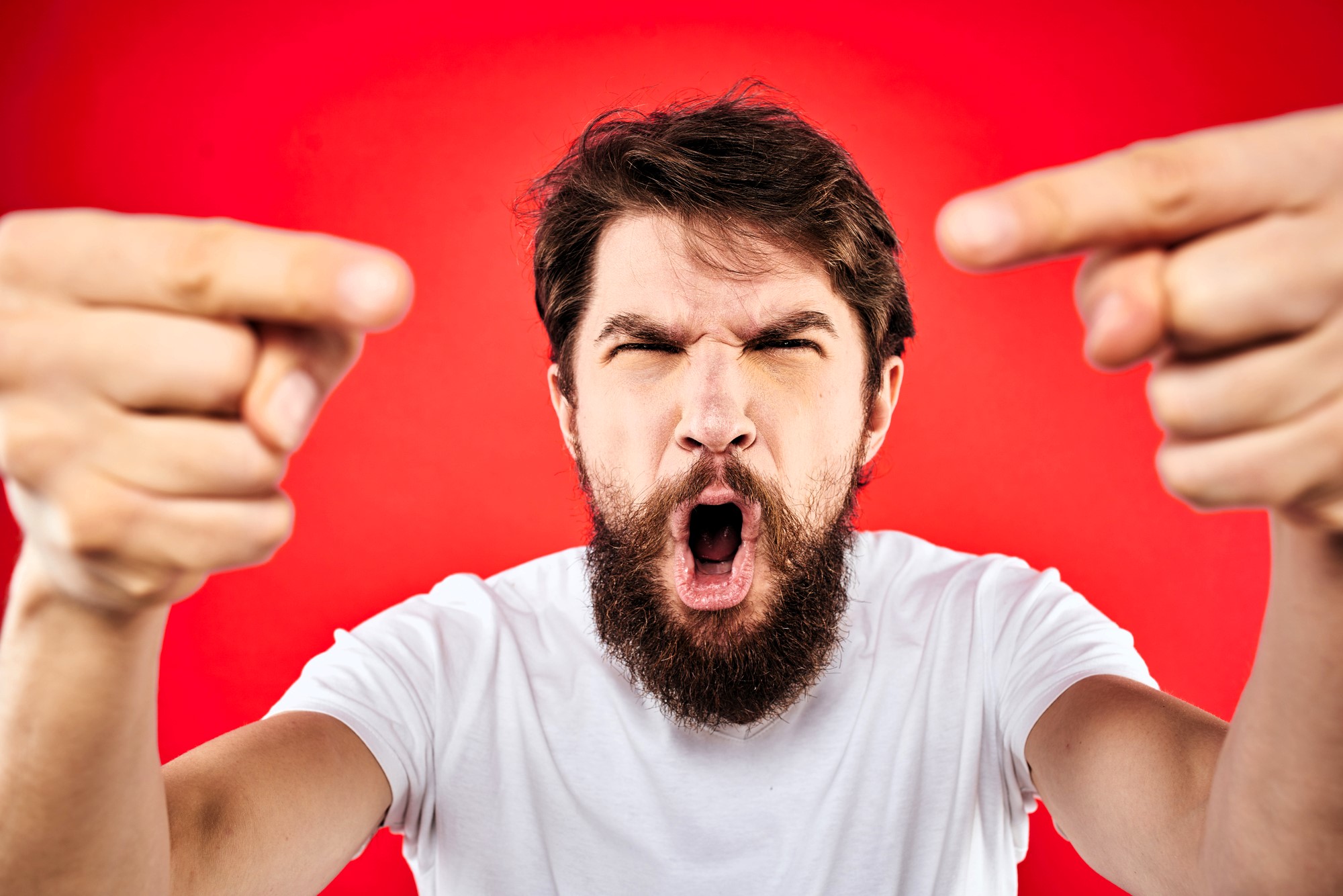 A bearded man in a white t-shirt, with an intense expression, points forcefully towards the camera. He is against a bright red background.