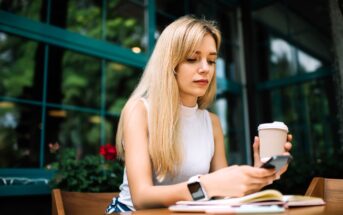 A woman with long blonde hair sits at a table holding a coffee cup and looking at her smartphone. She wears a white sleeveless top and a smartwatch. A notebook is on the table, and large windows with green frames are behind her.