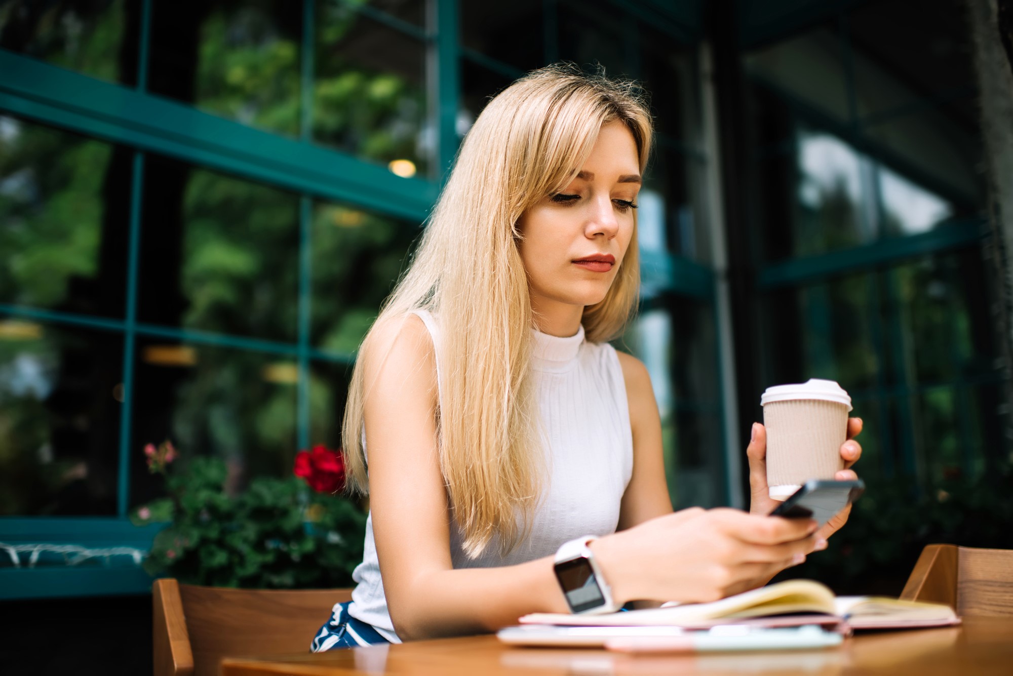 A woman with long blonde hair sits at a table holding a coffee cup and looking at her smartphone. She wears a white sleeveless top and a smartwatch. A notebook is on the table, and large windows with green frames are behind her.