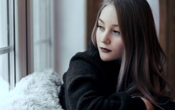 Woman with long hair and dark lipstick gazes thoughtfully out a window. She is wearing a dark, fuzzy sweater and is resting on a soft, textured surface. The lighting is soft and natural, creating a calm and introspective mood.