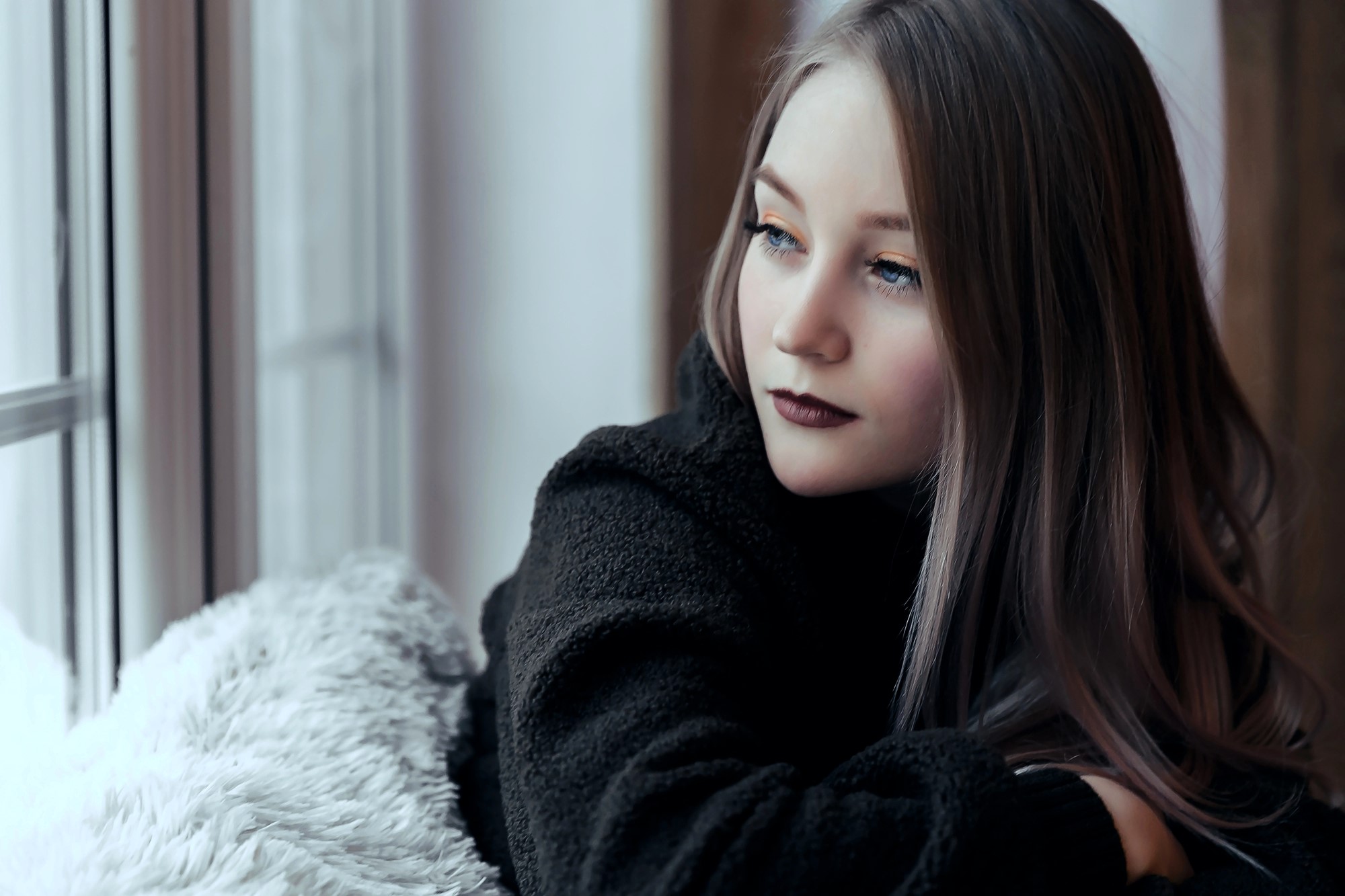 Woman with long hair and dark lipstick gazes thoughtfully out a window. She is wearing a dark, fuzzy sweater and is resting on a soft, textured surface. The lighting is soft and natural, creating a calm and introspective mood.