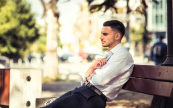 A man in a white shirt and dark pants sits on a wooden bench outdoors. He appears thoughtful, with his arms crossed, looking into the distance. Blurred trees and urban scenery are in the background.