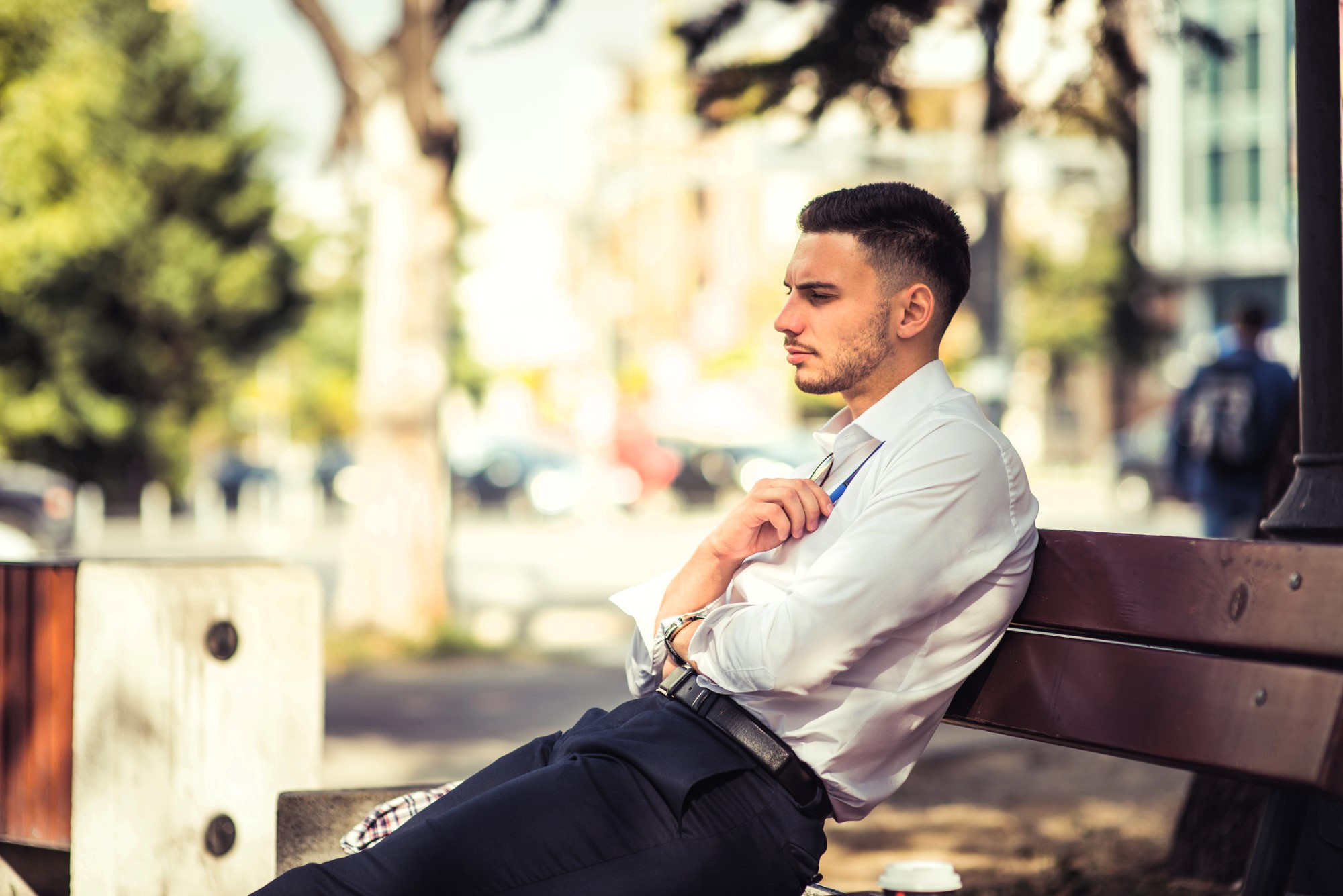 A man in a white shirt and dark pants sits on a wooden bench outdoors. He appears thoughtful, with his arms crossed, looking into the distance. Blurred trees and urban scenery are in the background.