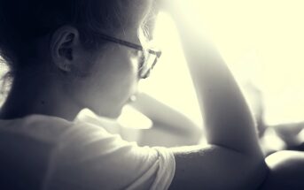 Black and white photo of a person in profile, wearing glasses and a white shirt. Their hand rests on their head, and they are looking towards a bright light, creating a soft, reflective mood.